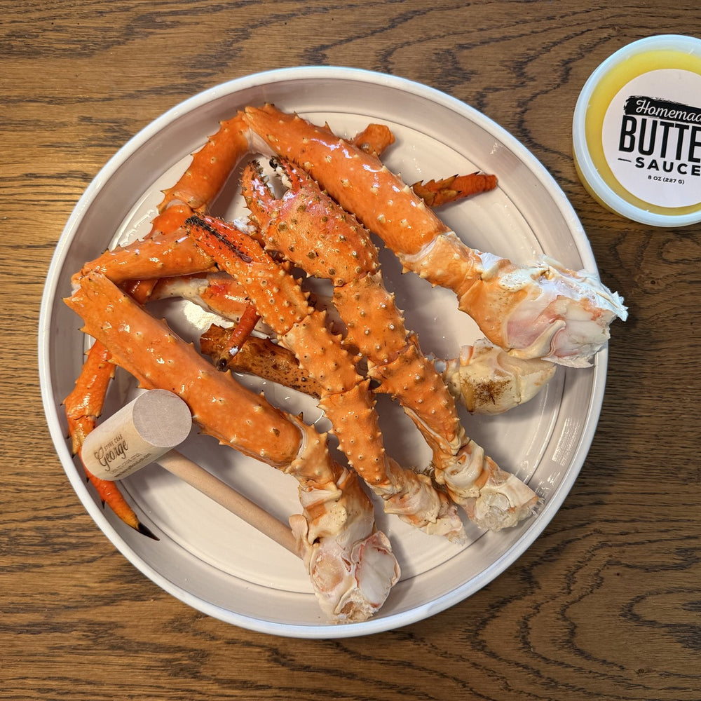 Image of Mega Colossal Alaskan King Crab Legs (6/9) - White plate with alaskan king crab legs and a container of butter sauce on a wooden surface.