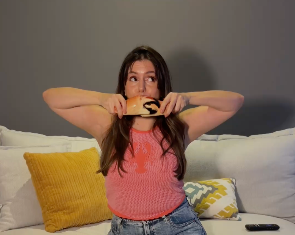 A woman sitting on a white couch holds a slice of bread with a black spider graphic across it, wearing a pink knit top and jeans in a living room setting.