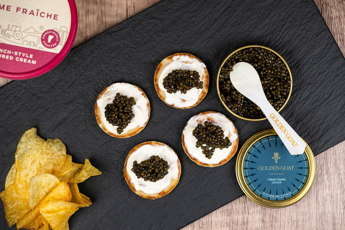 Caviar on crackers with chips and Golden Goat caviar containers on a black slate board.