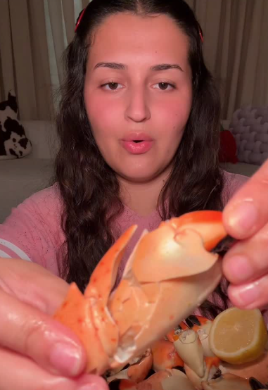 A close-up lifestyle shot of a woman holding a cooked crab claw with a lemon wedge, with a pile of crab claws in front of her.